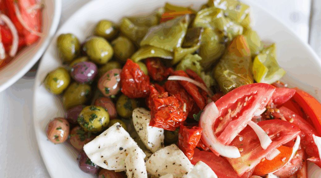 Image shows a plate of kalamata olives, pepperocinis, mozzarella, red onions, and tomatoes diced up and drizzled in olive oil.
